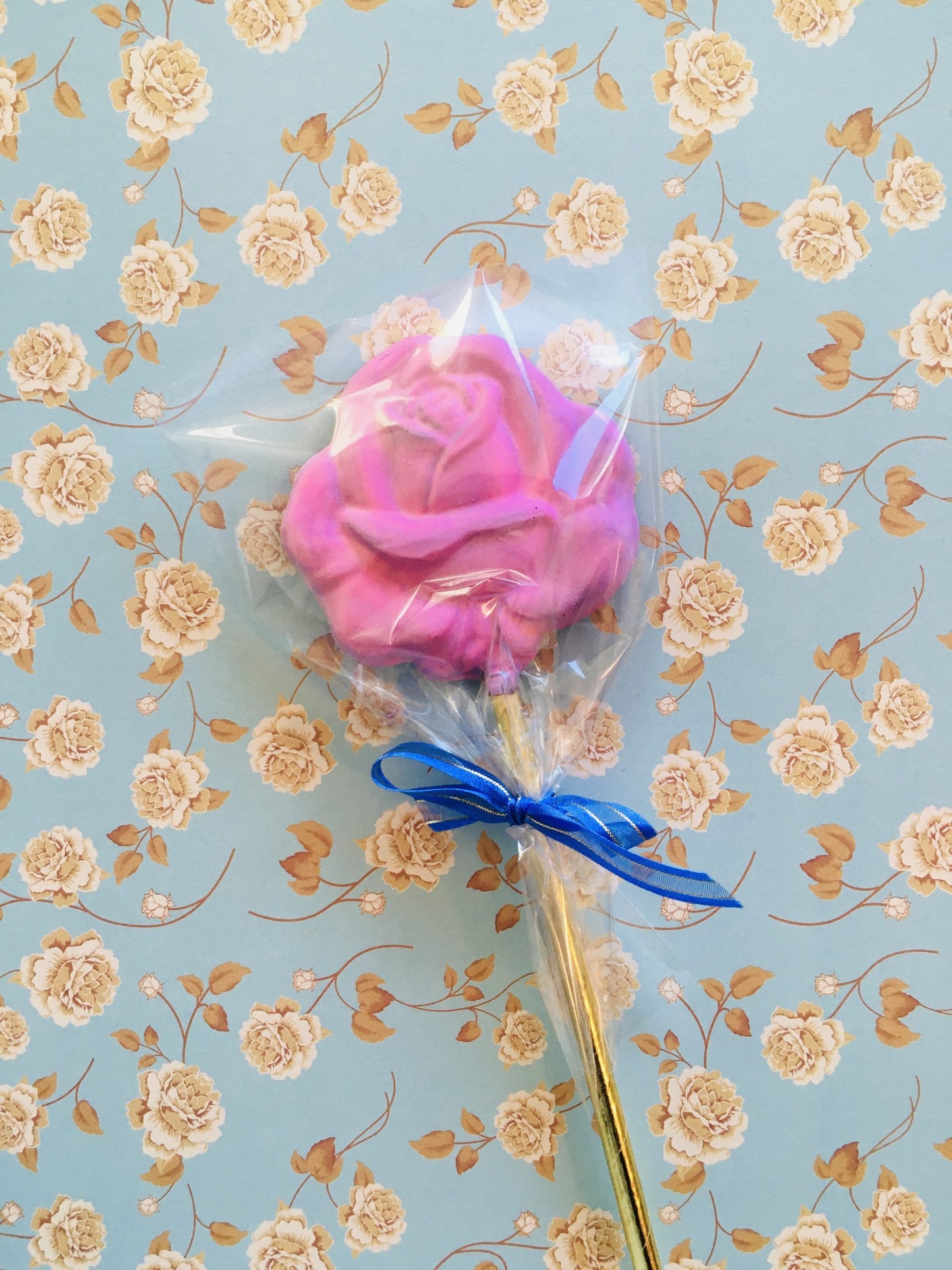 QUEST XO Rose Lolly: on a floral background, a lolly with a yellow stick, blue ribbon, and a rose lolly coloured pink.