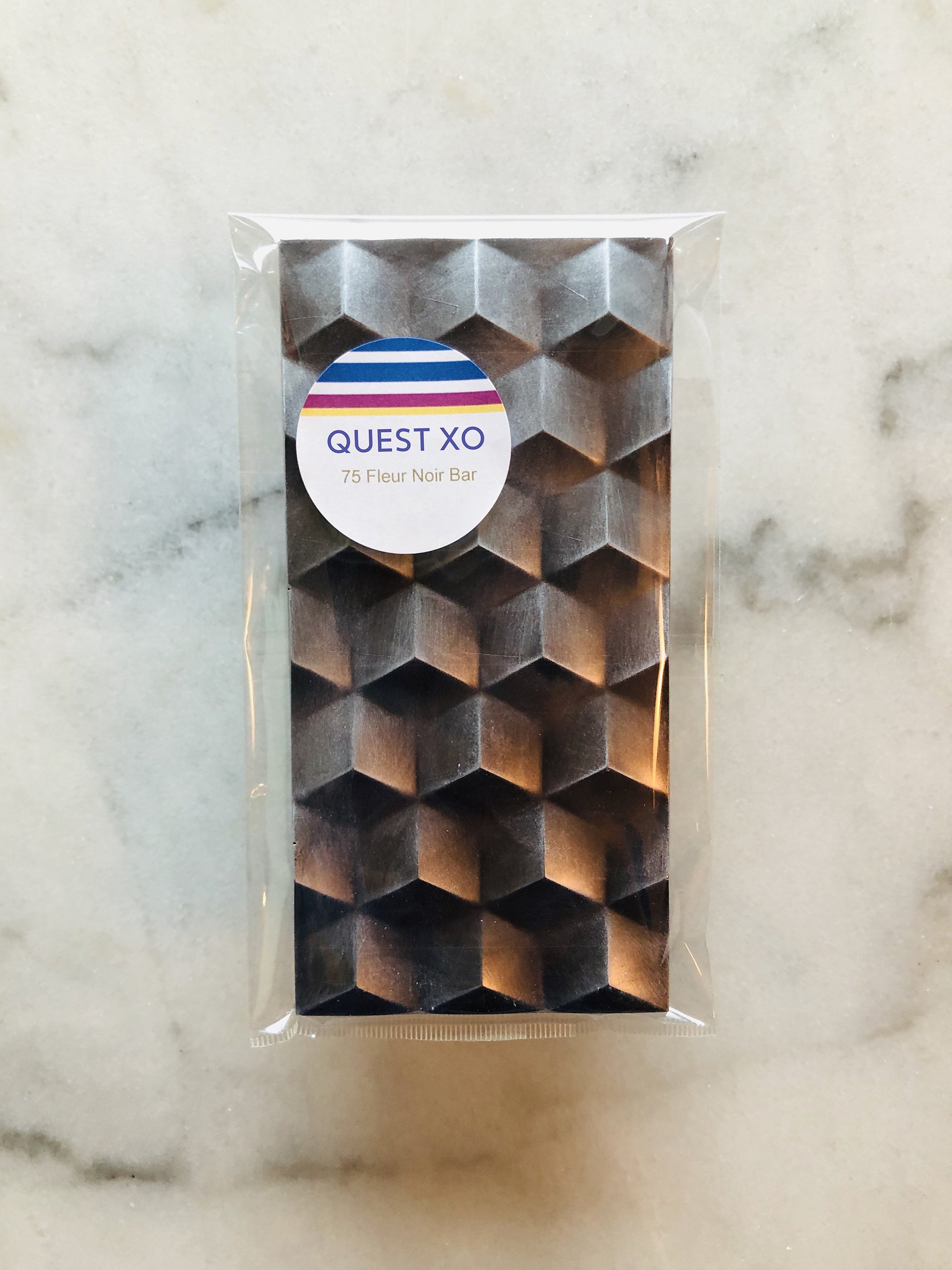 QUEST XO 75 Fleur Noir Bar: Dark brown chocolate bar with hexagonal shapes wrapped in a transparent plastic with QUEST XO logo placed on a marble background.
