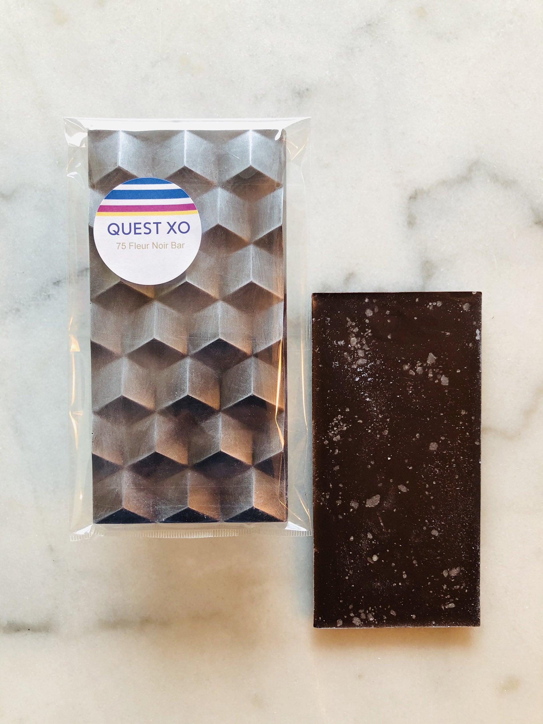 QUEST XO 75 Fleur Noir Bar: Two dark brown chocolate bars with hexagonal shapes wrapped in a transparent plastic with QUEST XO logo placed on a marble background.