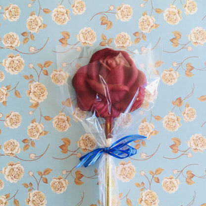 QUEST XO Rose Lolly: on a floral background, a lolly with a yellow stick, blue ribbon, and a rose lolly coloured maroon
