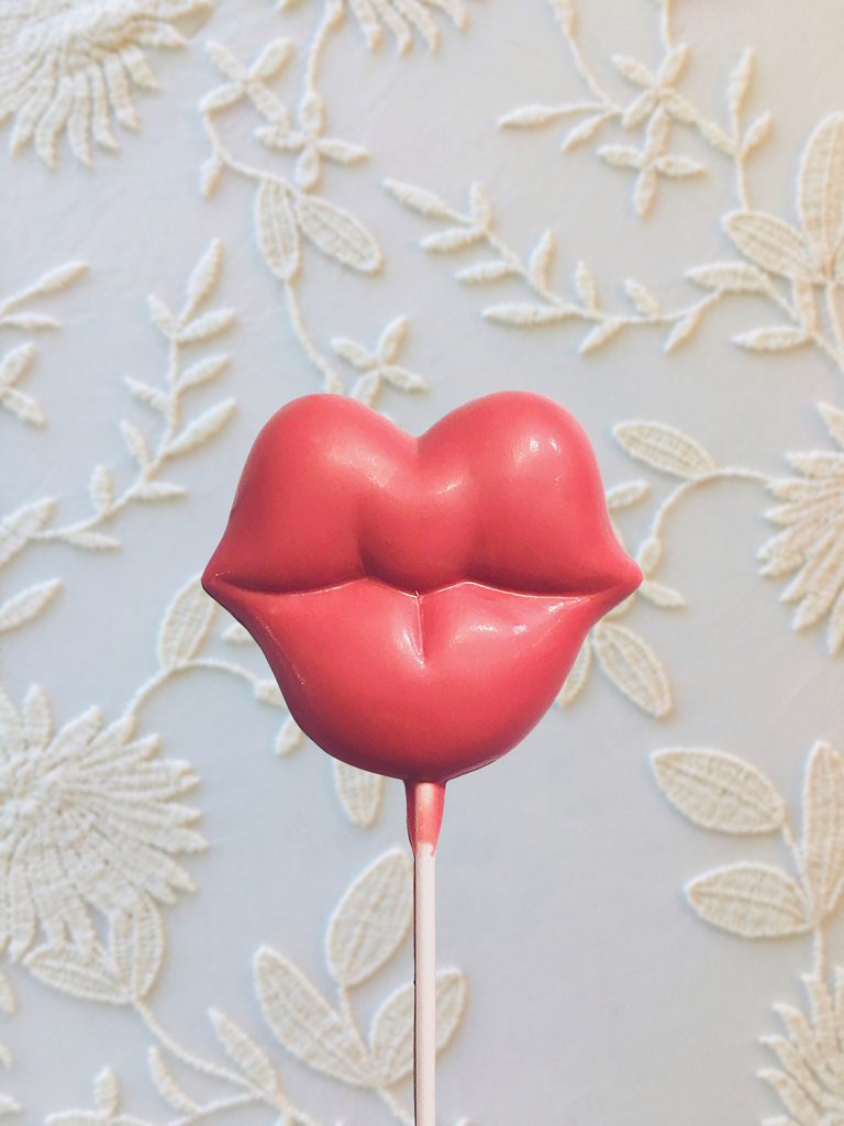 QUEST XO Kiss Lolly: Lip-shaped lolly on a wooden stick with floral background.