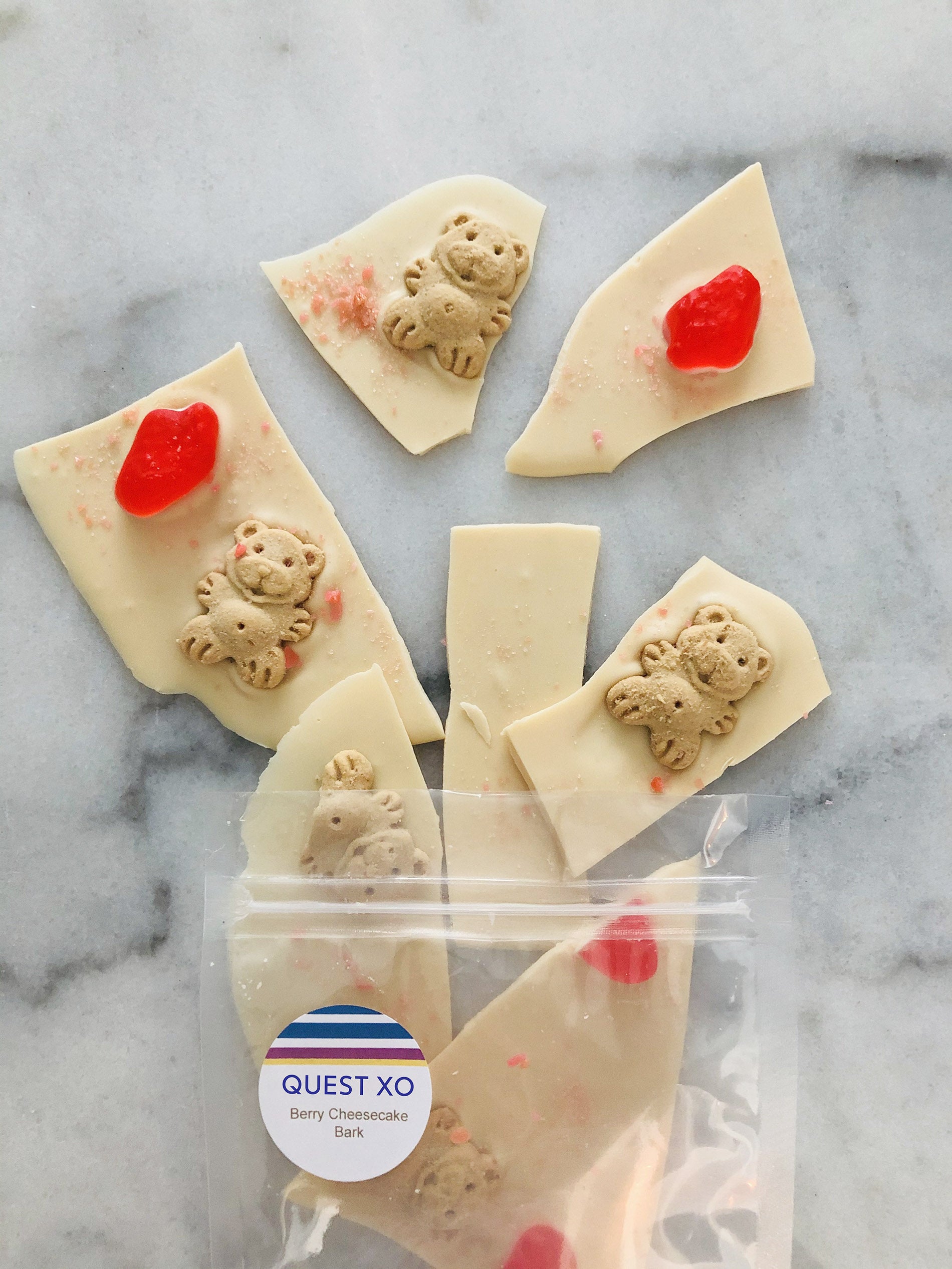 QUEST XO Berry Cheesecake Bark: Pieces of white chocolate with teddy bears on it falling out of a transparent pack on a marble background.