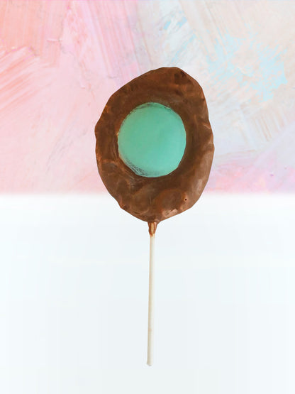QUEST XO Egg Lolly: Brown lolly with green coloured centre with white stick placed on a pink and white background.