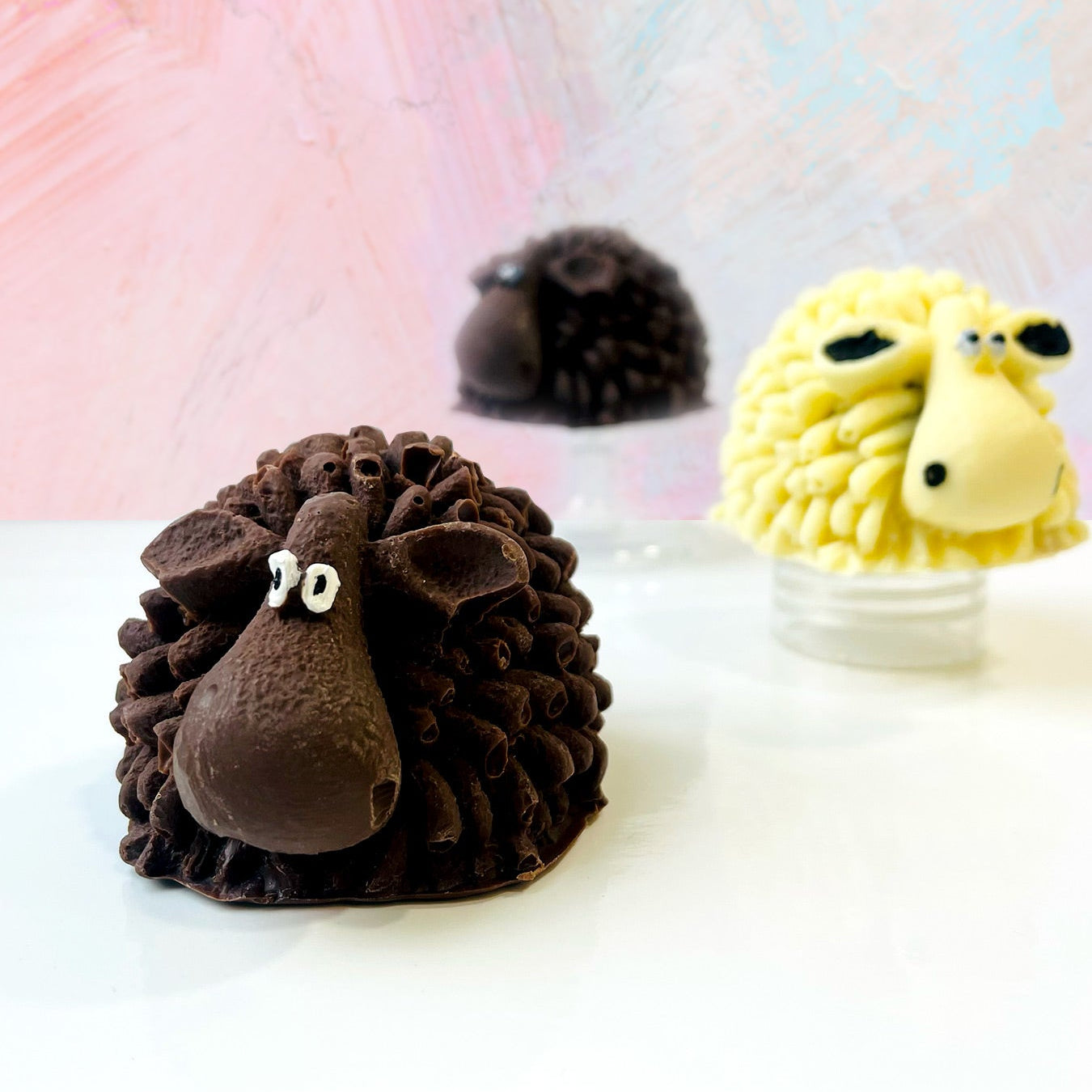 QUEST XO Sheepy Hot Cocoa Bomb: 3 chocolate sheep cocoa bombs. Right at the back, blurry brown sheep. Centre right is a yellow sheep. Foreground left is a brown sheep.