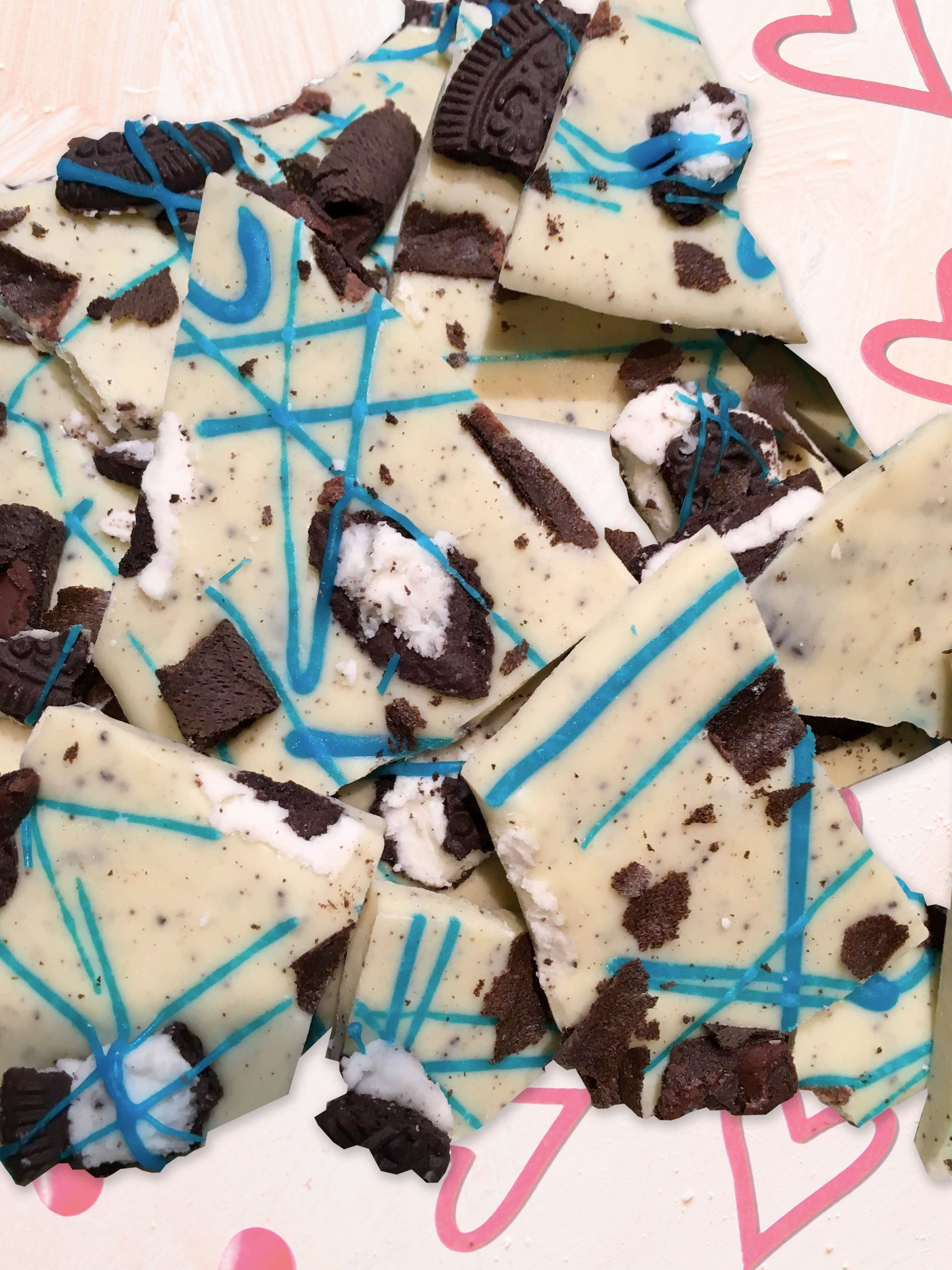 QUEST XO Cookies + Cream: Pieces of white chocolate decorated with Oreos and blue coloured chocolate placed on a background with pink hearts.